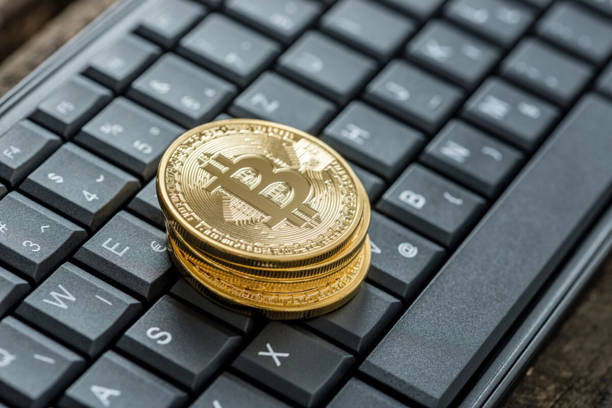 A stack of shiny gold Bitcoin coins rests on a black computer keyboard, symbolizing digital currency and online financial transactions.