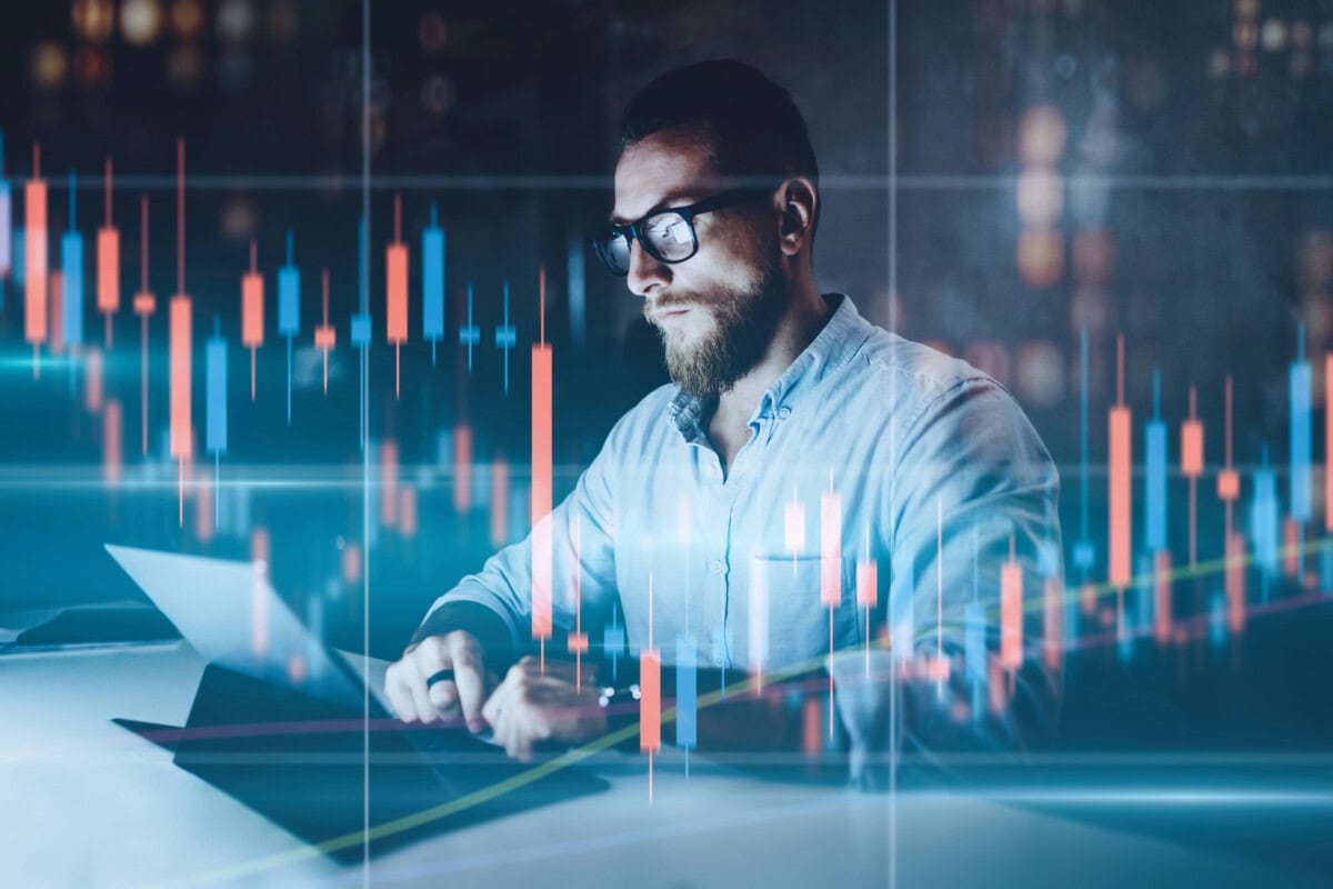 A man with glasses and a beard works on a laptop in a dimly lit room, with colorful financial graphs and candlestick charts overlaid in the foreground, suggesting financial analysis or trading.