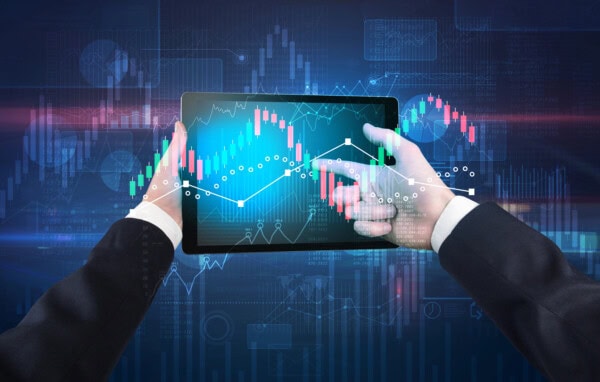A person in a suit holds a tablet displaying colorful financial charts and graphs, with digital financial data and analytics in the background.