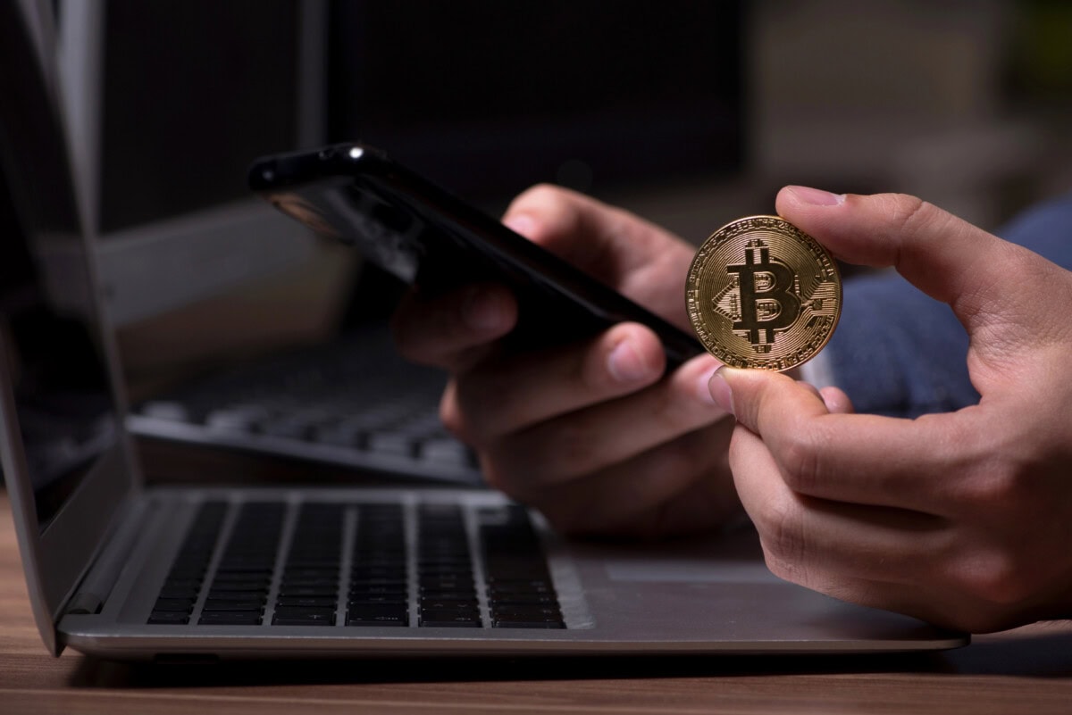A person holds a gold-colored Bitcoin coin in one hand and a smartphone in the other, with a laptop in the background on a desk.