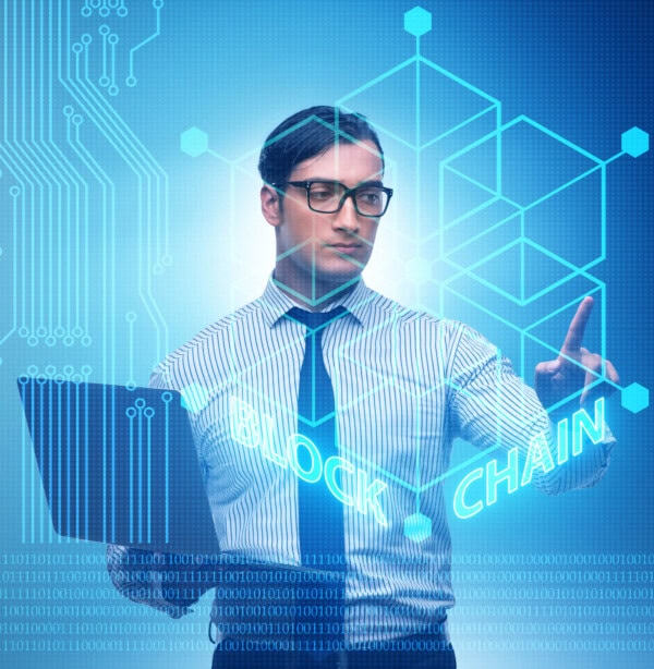 A man in business attire holding a laptop interacts with a virtual blockchain interface, surrounded by digital graphics, binary code, and the word “BLOCKCHAIN” in glowing letters.