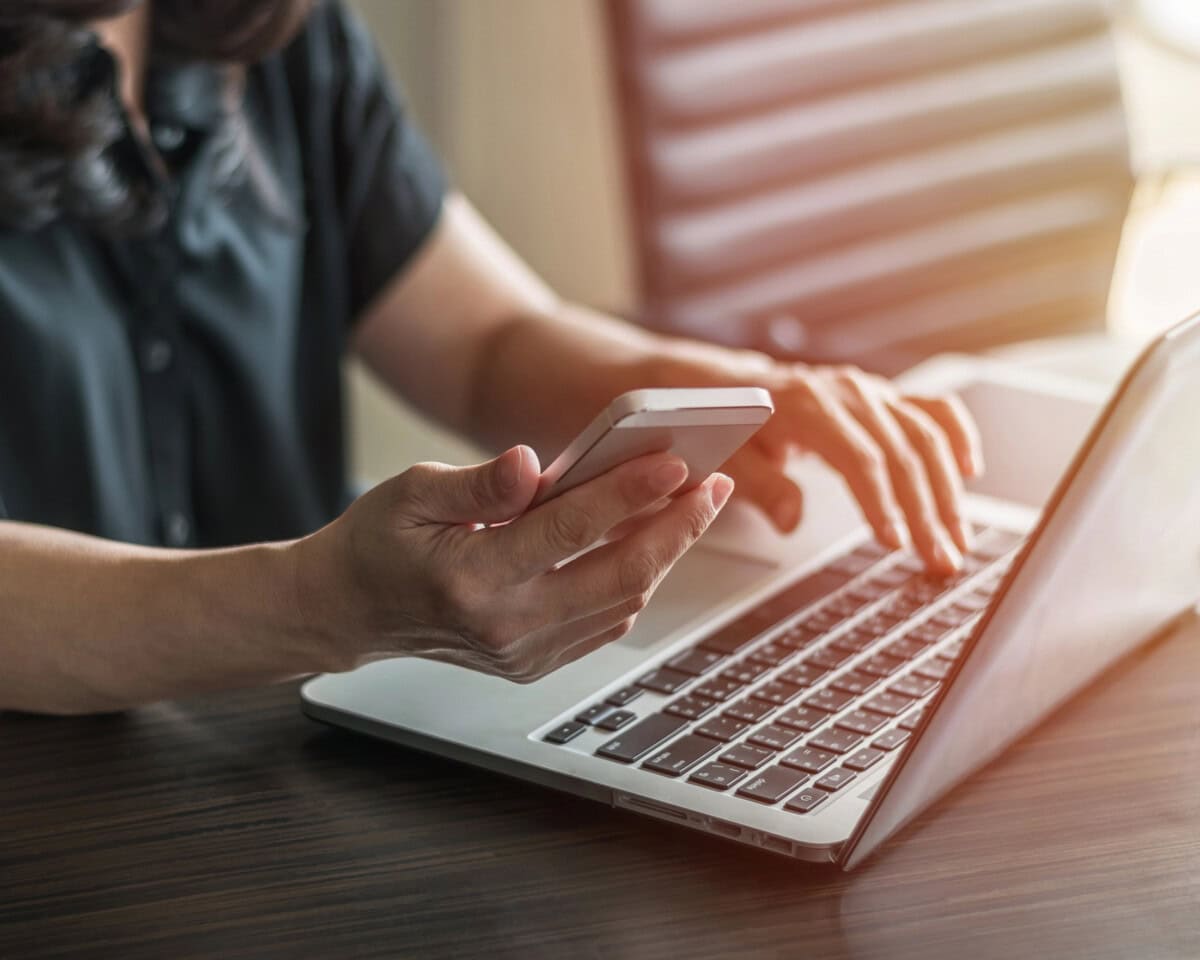 A person sitting at a desk using a smartphone in one hand and typing on a laptop keyboard with the other hand. The scene is well-lit, and the individual is wearing a dark short-sleeved shirt.