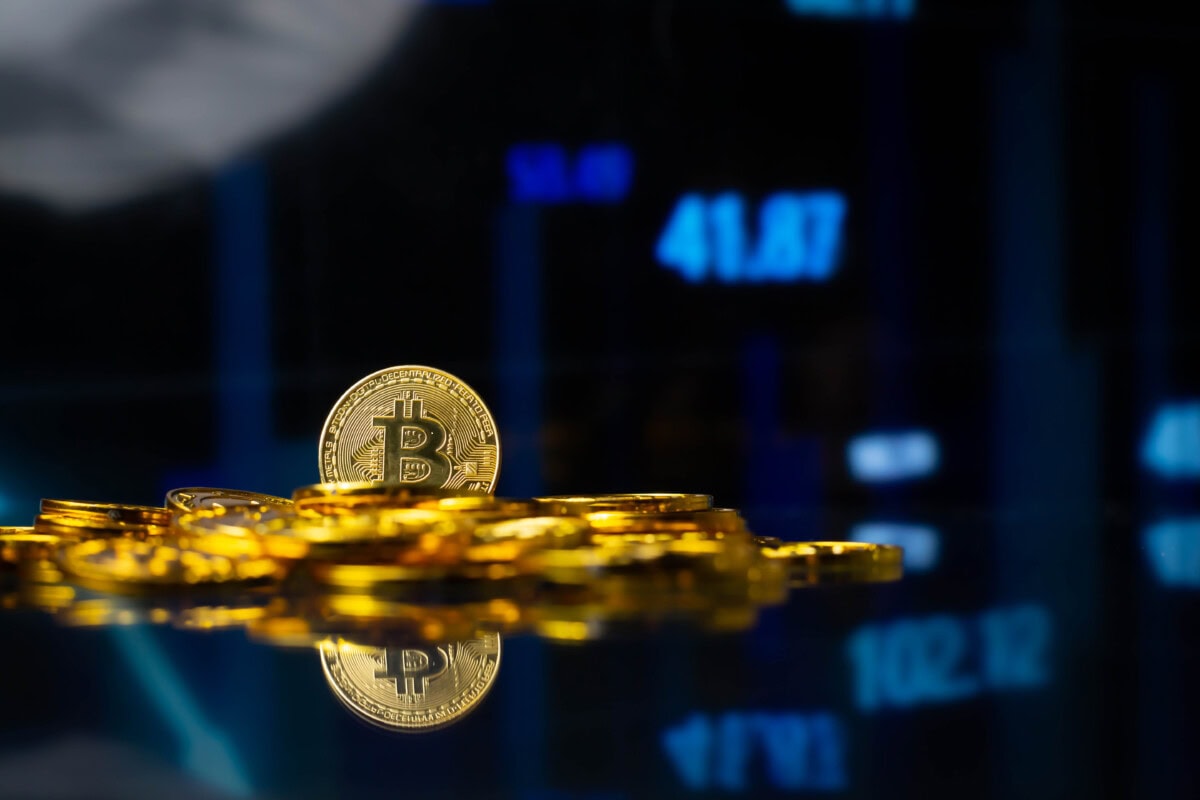 A stack of shiny gold Bitcoin coins sits on a reflective surface, with a blurred digital stock market chart displaying blue numbers in the background.