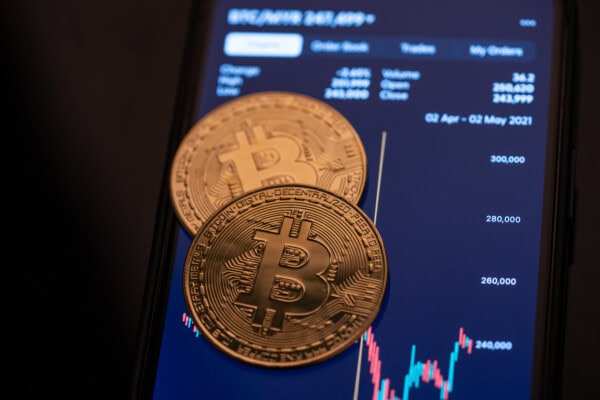 Two gold-colored physical Bitcoin coins rest on a smartphone screen displaying a cryptocurrency price chart and trading data.