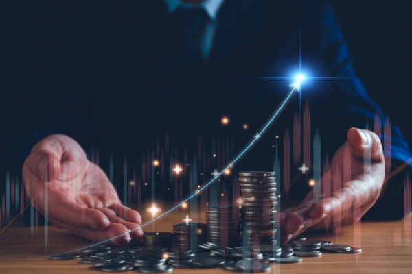 A person in a suit holds out their hands around stacks of coins on a table, with a glowing upward arrow and financial graphs overlayed, symbolizing financial growth and success.
