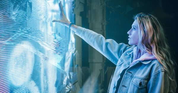 A woman in a denim jacket touches a glowing, futuristic light wall, illuminating her face with blue light.