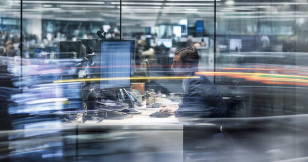 A man works at a computer in a modern office with glass walls, surrounded by blurred motion of lights and people.