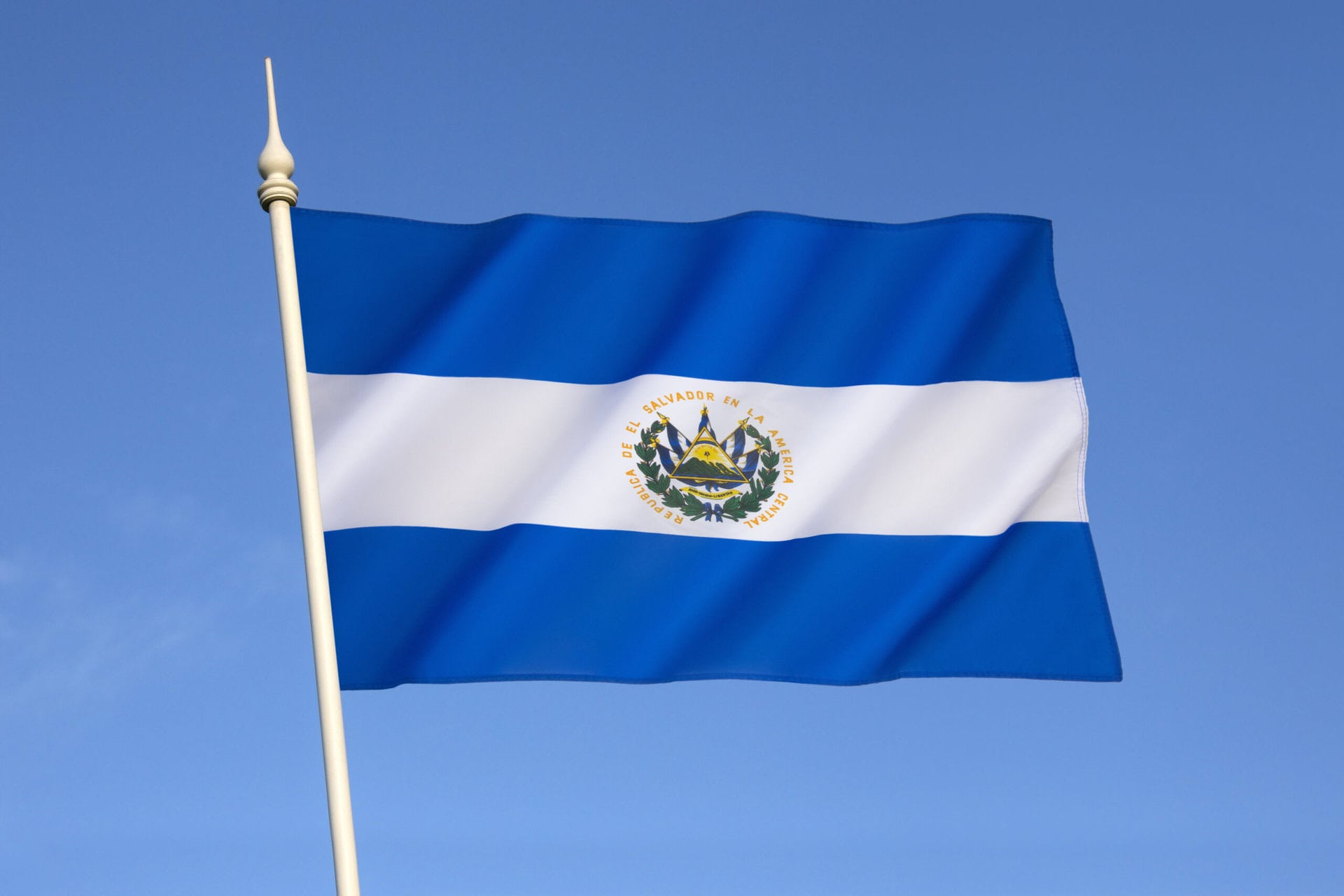 The flag of El Salvador with blue and white horizontal stripes and the national coat of arms in the center, waving on a flagpole against a clear blue sky.