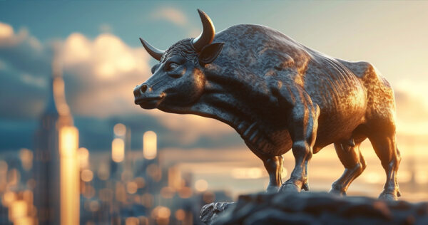 A bronze bull statue stands on a rock ledge with a city skyline and cloudy sky in the background at sunset.