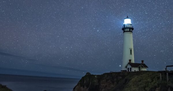 A lighthouse stands on a cliff by the sea, its light shining under a clear night sky filled with stars.