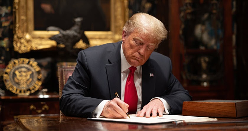 A man in a suit and red tie sits at a wooden desk, writing on a document with a pen. There are official-looking items and a framed painting in the background.