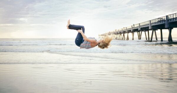 A person performs a backflip on the beach near the shoreline, with a pier extending into the ocean under a cloudy sky.