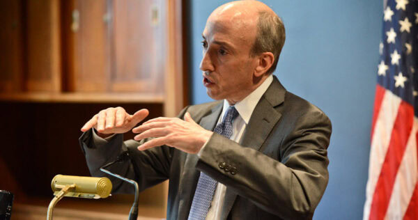 A man in a suit gestures with his hands while speaking at a podium, with an American flag and wooden shelves in the background.