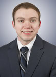 A man with short brown hair wearing a dark suit, white shirt, and striped tie poses for a formal portrait against a light gray background.