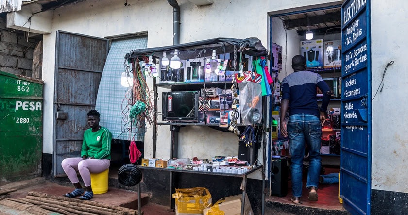 A small roadside electronics shop displays cables, bulbs, and gadgets; a man works inside while a woman sits outside on a yellow container.