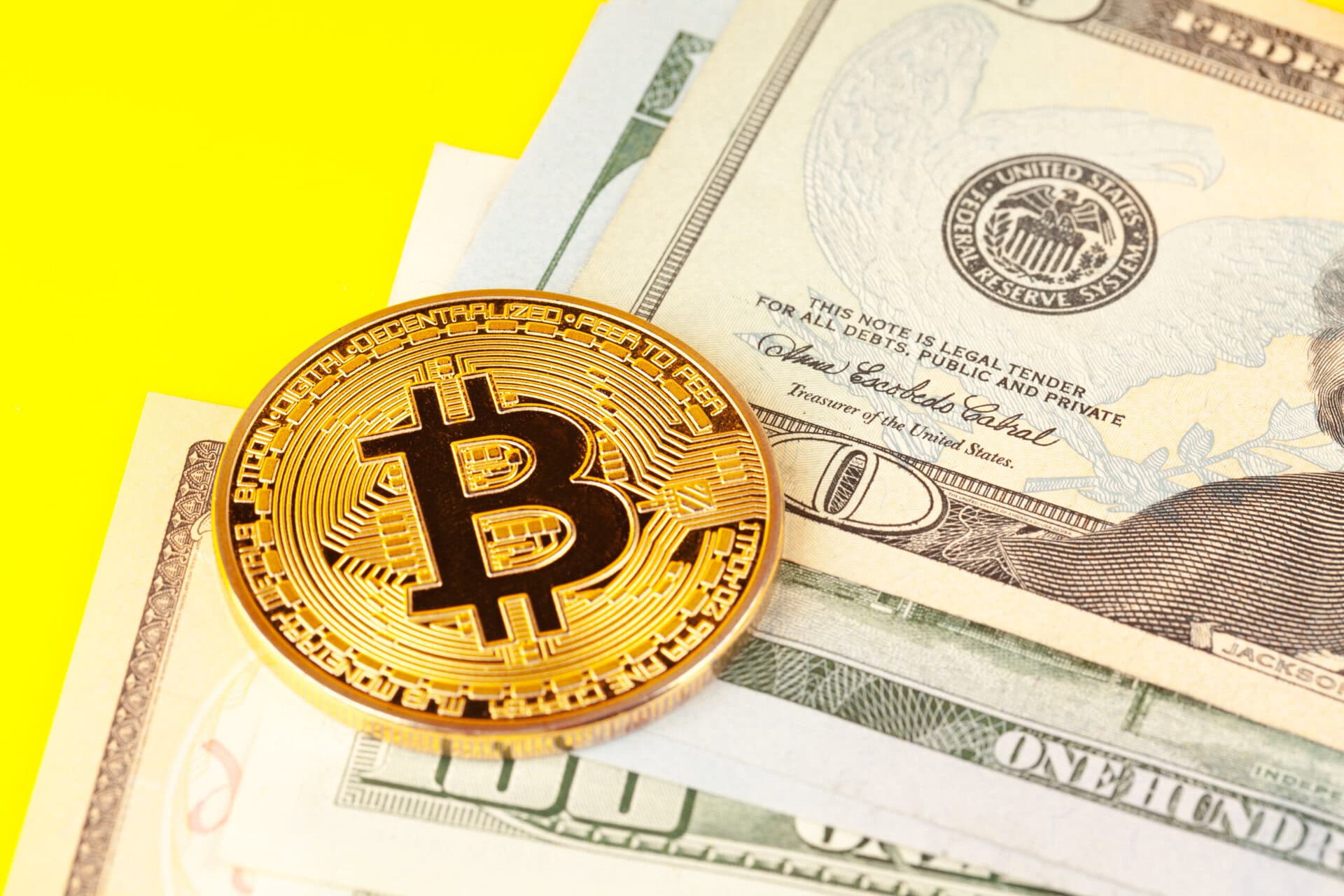 A gold Bitcoin coin placed on top of several U.S. dollar bills, including a visible one hundred dollar bill, against a bright yellow background.