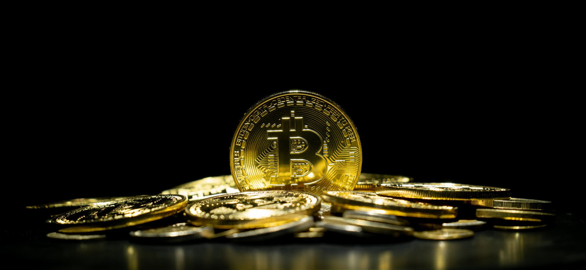Several gold-colored Bitcoin tokens are stacked and scattered on a dark surface with a black background.