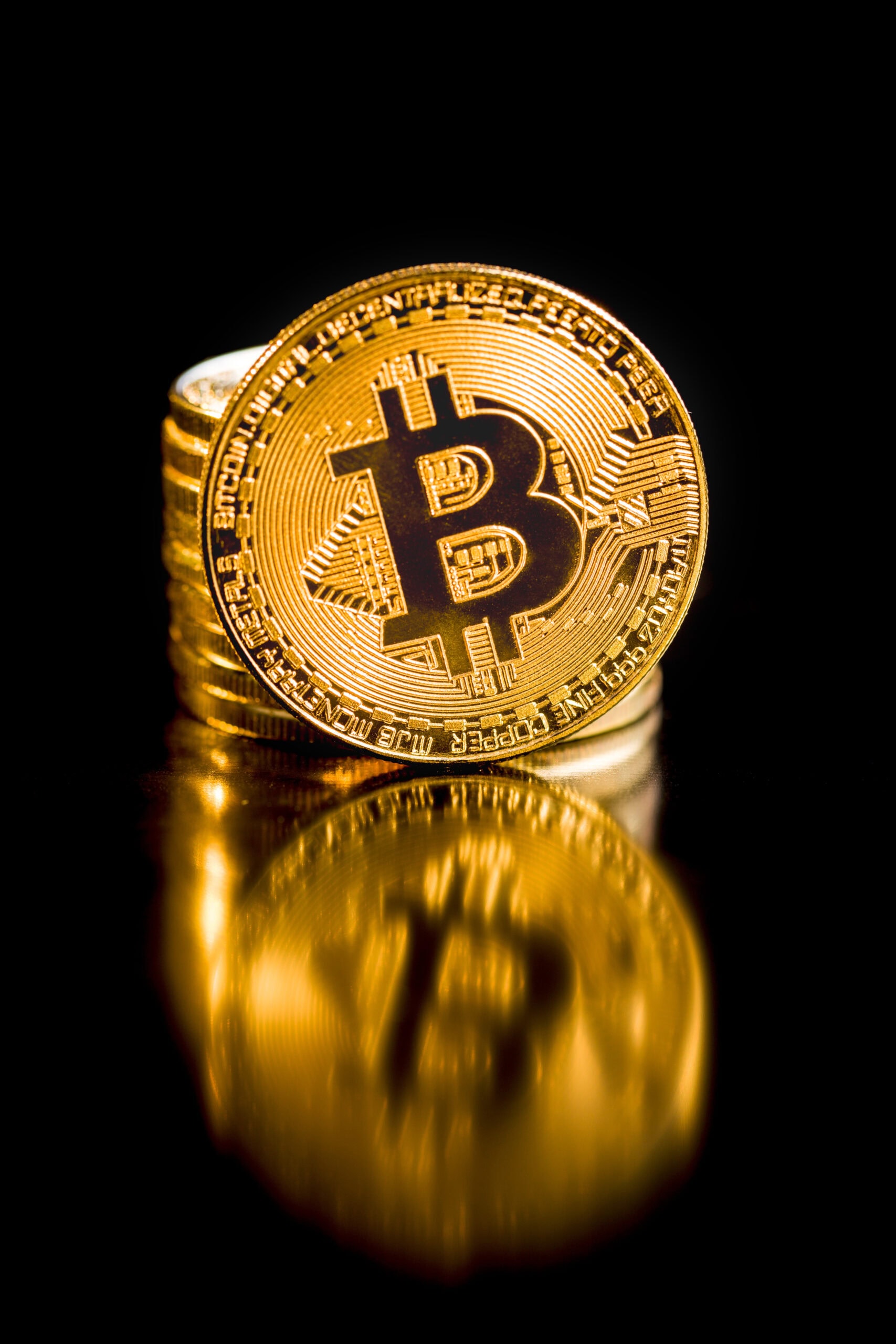 A stack of shiny gold-colored coins with the Bitcoin symbol, reflecting on a black surface. The focus is on the front coin, displaying the Bitcoin logo prominently.