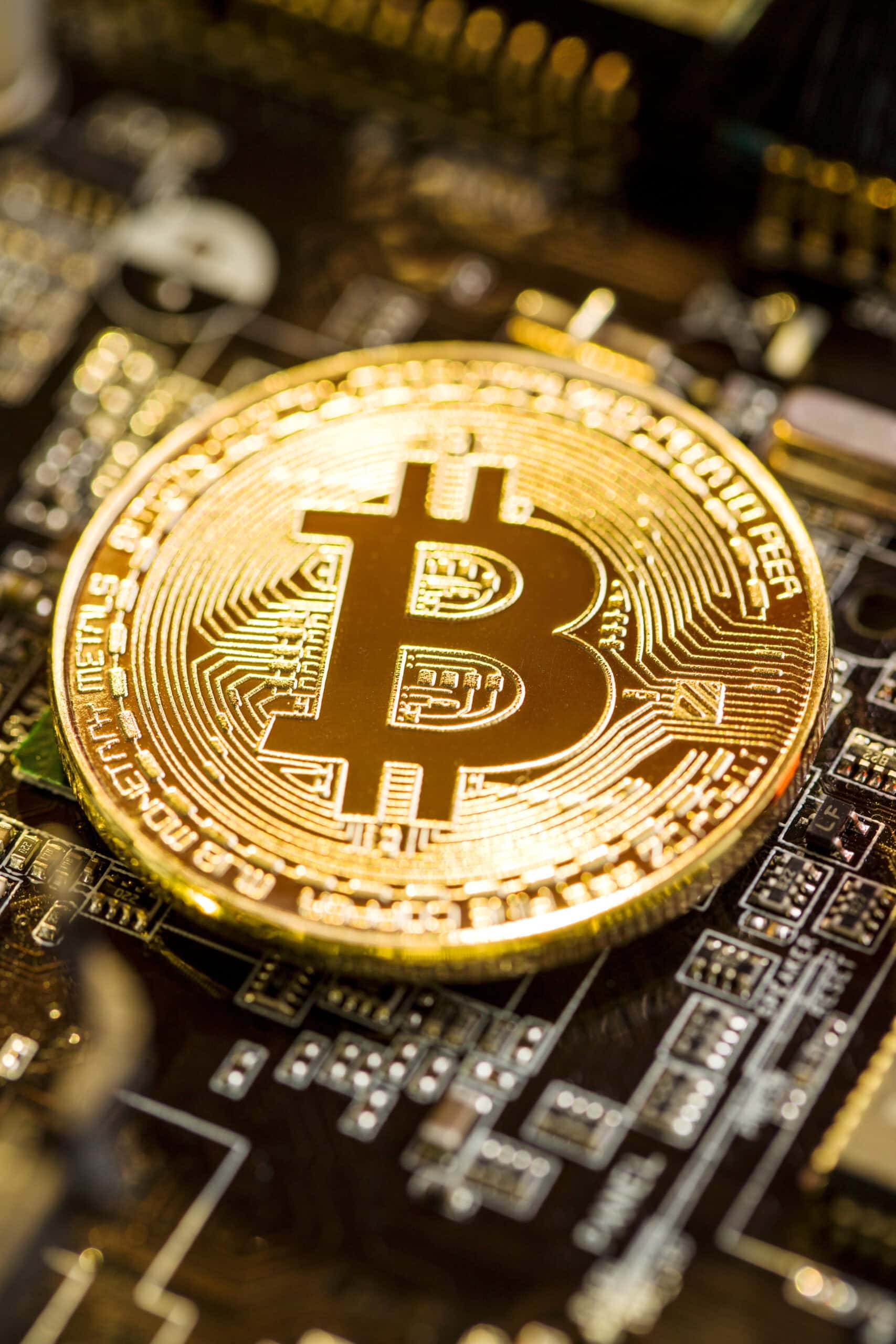 A shiny gold-colored physical Bitcoin coin rests on top of a computer circuit board, symbolizing the connection between cryptocurrency and digital technology.