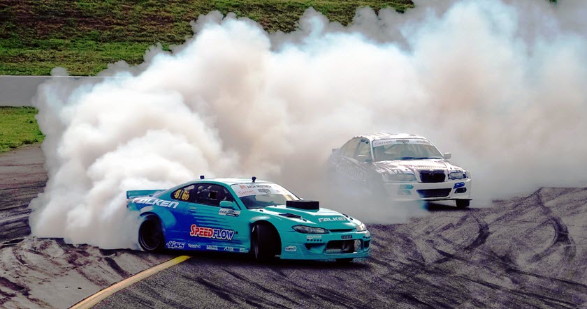 Two cars drift around a curve on a racetrack, producing large clouds of smoke from their tires.