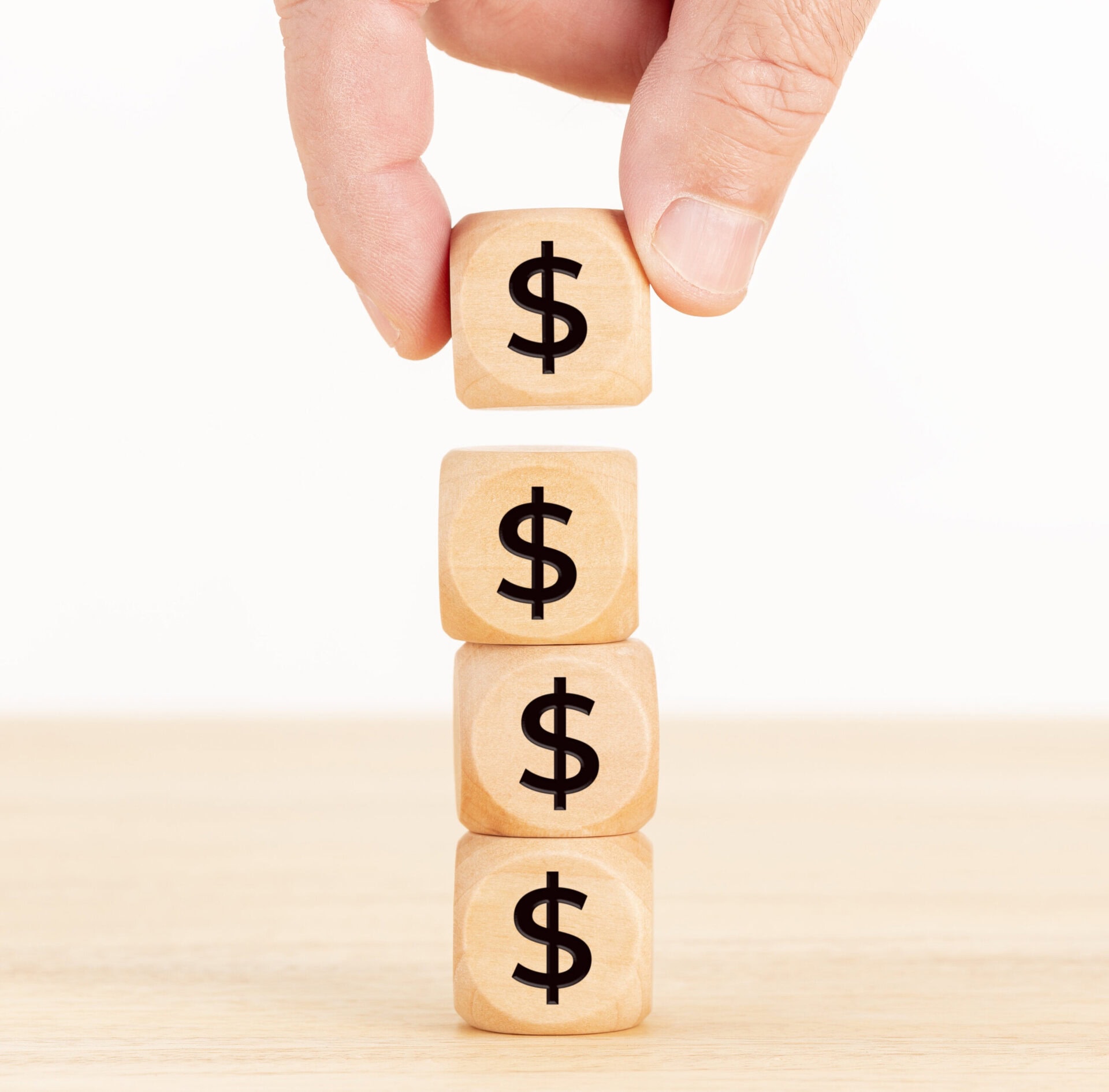 A hand stacks a wooden block with a dollar sign on top of three other similar blocks, all displaying dollar signs, on a light wooden surface.