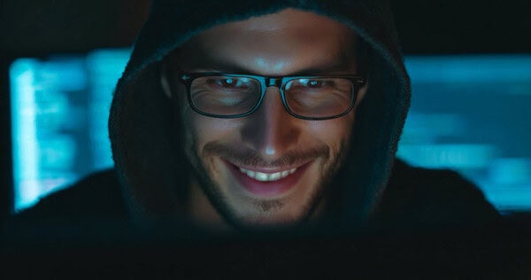 A man wearing glasses and a hooded sweatshirt smiles while looking at a computer screen, with blue light from the monitor illuminating his face in a dark room.