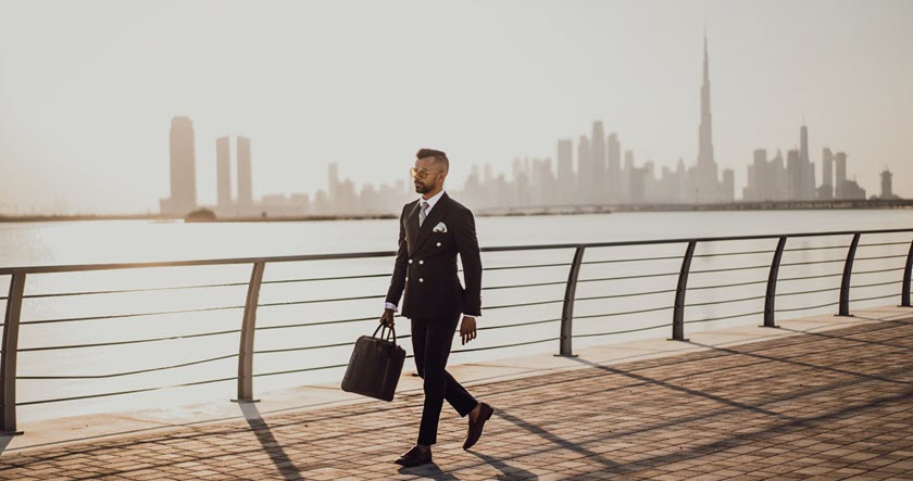 A man in a black suit and sunglasses walks along a waterfront promenade with a city skyline in the background, carrying a briefcase.