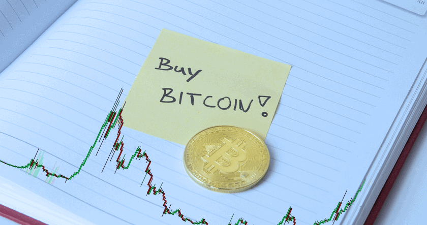 A gold Bitcoin coin and a sticky note with "Buy BITCOIN!" written on it are placed on an open notebook, with a cryptocurrency price chart in the background.