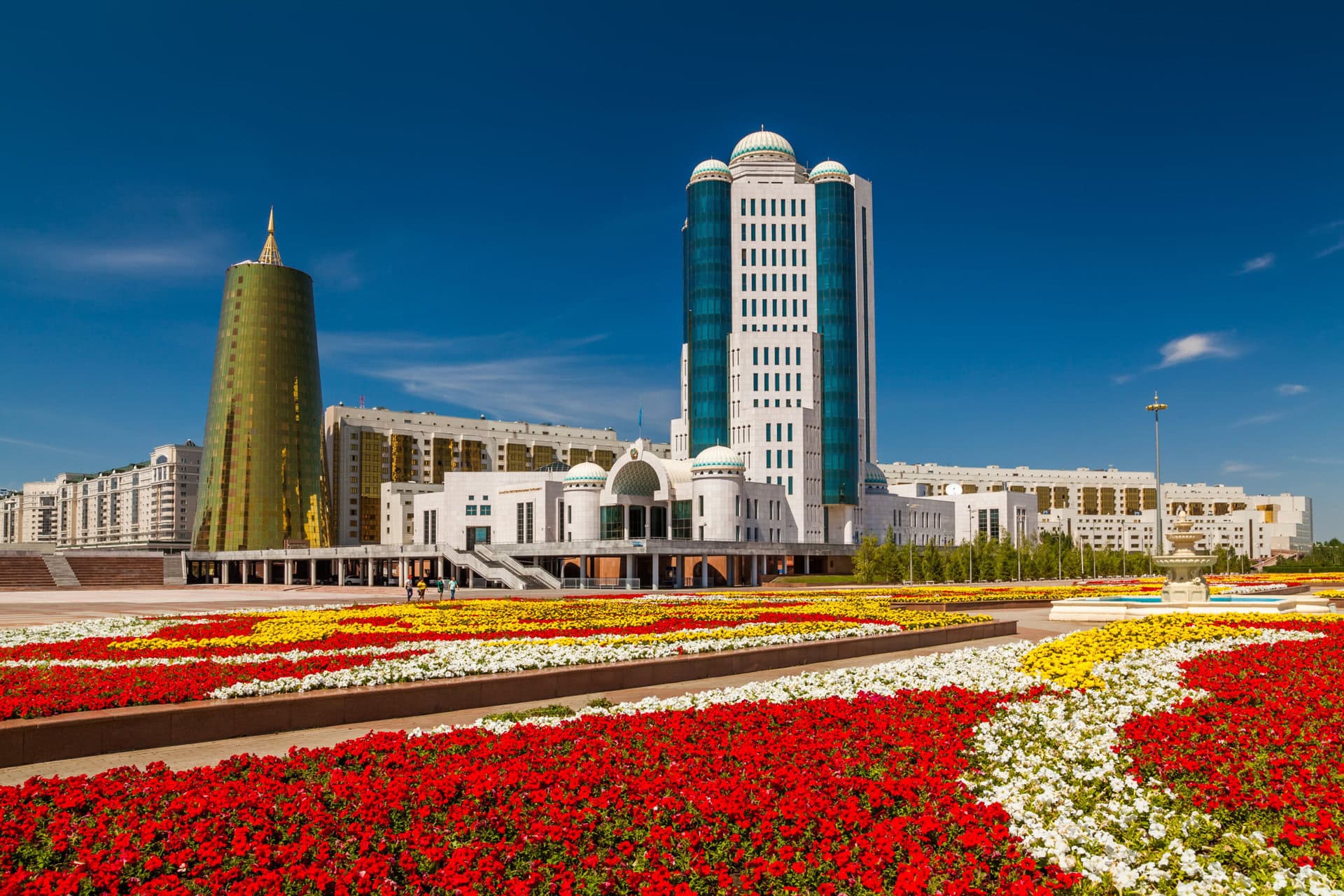 Modern buildings with unique architecture stand behind vibrant red, white, and yellow flower beds under a clear blue sky.