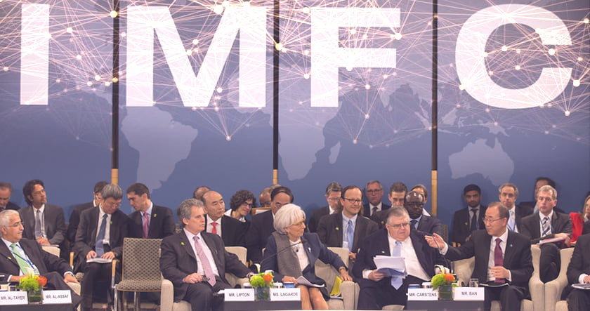 A group of officials sit and discuss on stage at an IMF conference, with a world map and "IMF" letters in the background.
