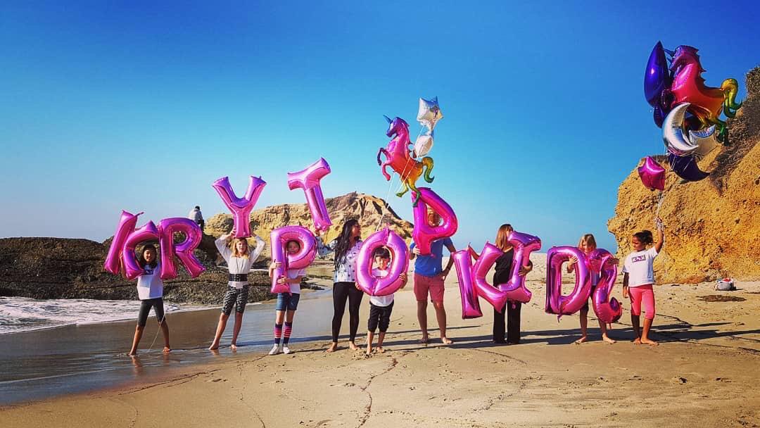 A group of children and adults stand on a beach holding pink balloons that spell "KRYPTOPOKIDS," with colorful star and unicorn balloons nearby.