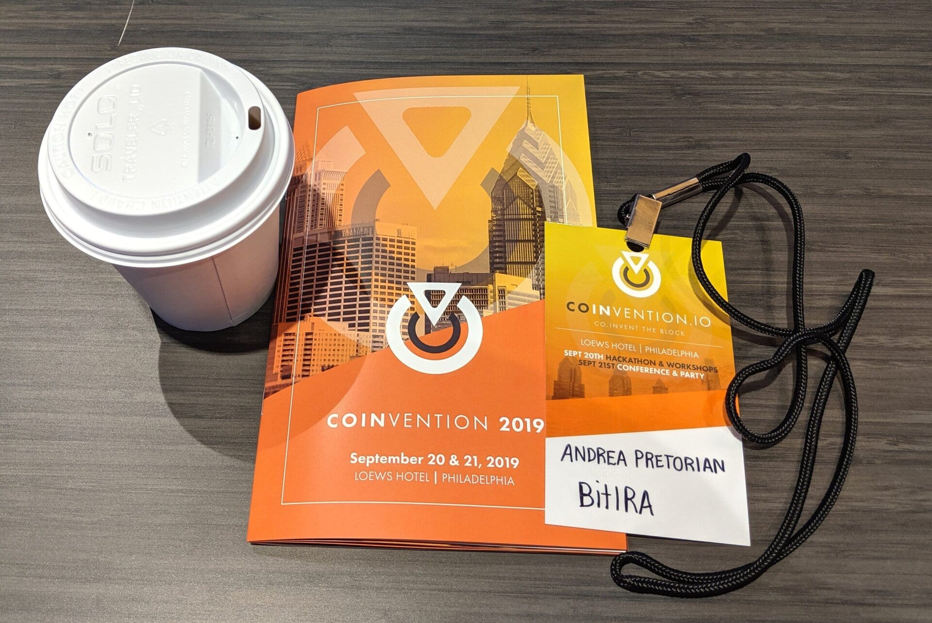 A coffee cup, event program, and a name badge for Coinvention 2019 are on a table. The badge displays the name Andrea Pretorian and the word “Bithira.”.