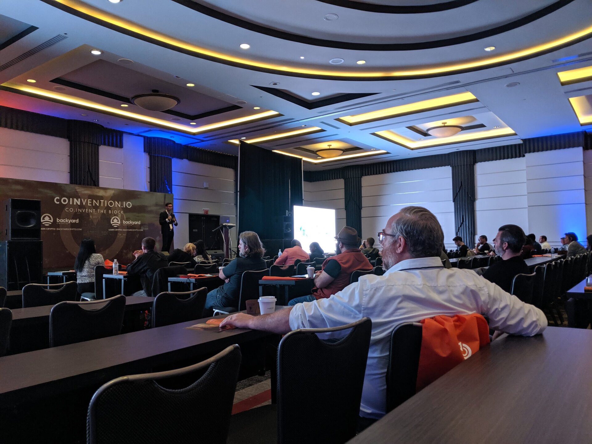 Audience seated in a conference room watching a speaker present on stage at COINVENTION.IO event, with a large screen and banners visible.