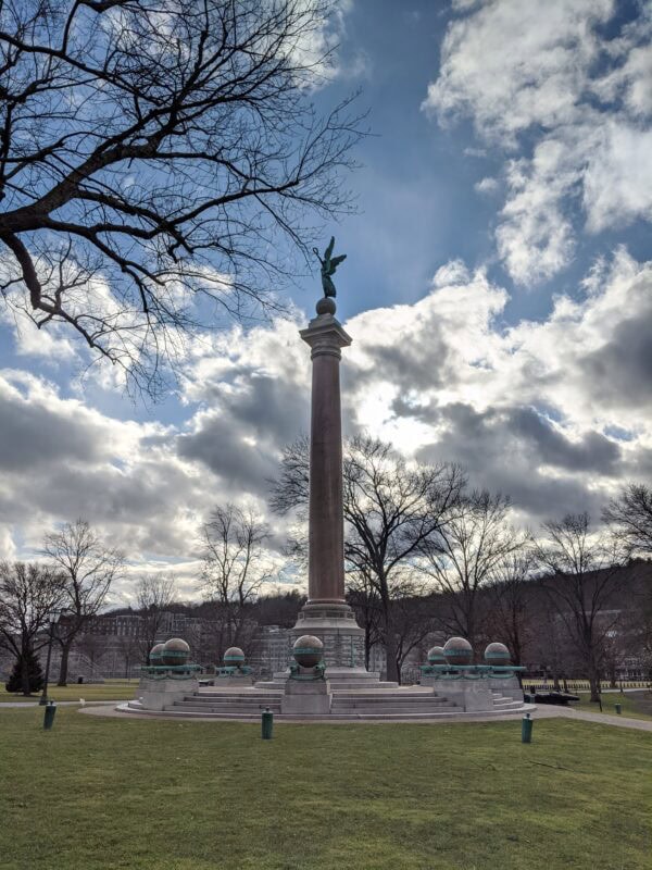 A tall column monument topped with a winged statue stands in a park surrounded by bare trees and a cloudy sky.