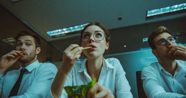 Three young adults in business attire sit indoors, closely side by side, eating snacks and looking attentively in the same direction, as if watching a presentation or event.