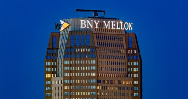A high-rise building at dusk features the illuminated BNY Mellon logo and sign near its top against a clear blue sky.