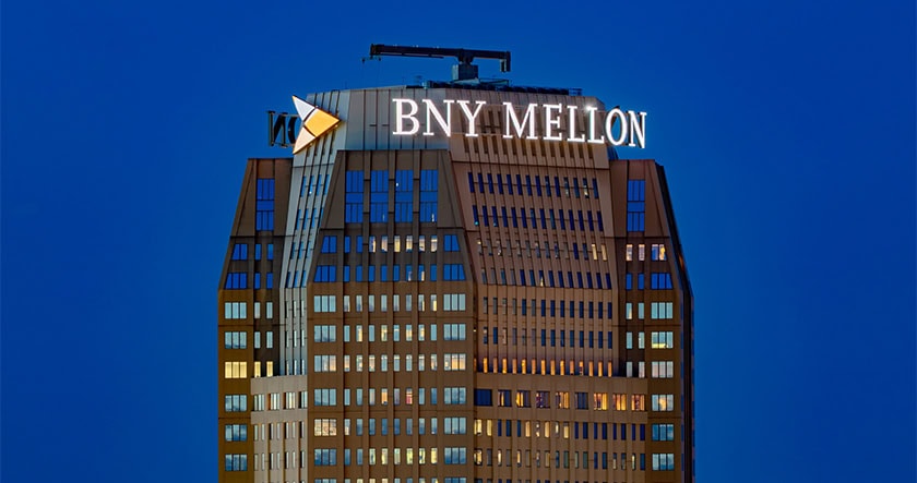 A high-rise building at dusk features the illuminated BNY Mellon logo and sign near its top against a clear blue sky.