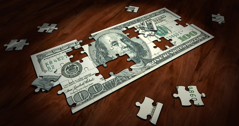 A $100 bill is shown as a jigsaw puzzle with several pieces missing, lying on a wooden surface.