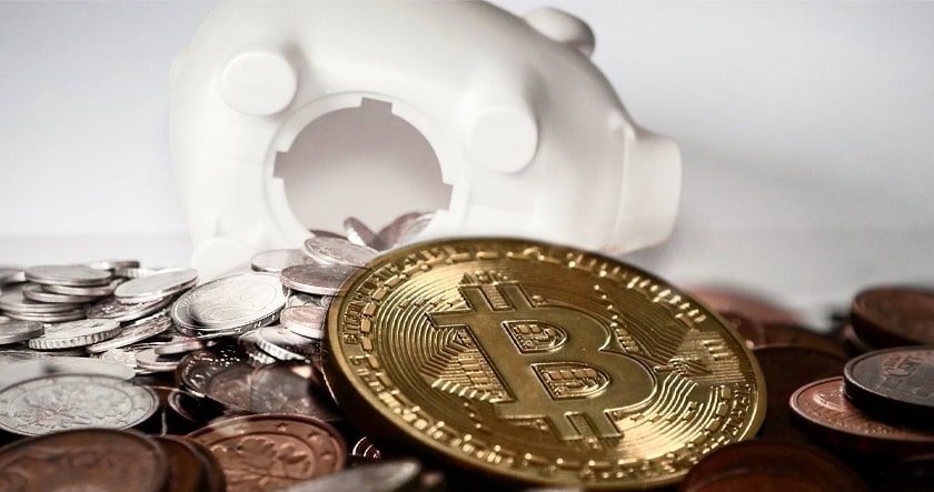 A gold Bitcoin token lies among assorted coins in front of an empty white piggy bank.