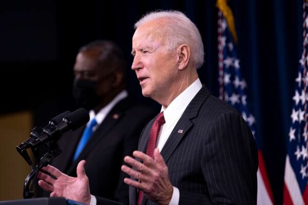 A man in a suit and red tie speaks at a podium with microphones, with American flags and another person in the background.