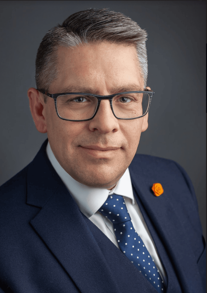 A man wearing glasses, a dark blue suit, a white shirt, and a blue polka dot tie with a small orange pin, poses against a plain grey background.