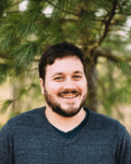A man with dark hair and a beard, wearing a dark sweater, smiles while standing outdoors in front of green pine tree branches.