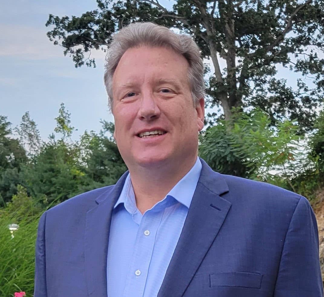 A man with light hair wearing a blue suit jacket and light blue shirt stands outdoors, smiling, with green trees and a clear sky in the background.