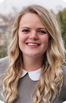 A young woman with long, wavy blonde hair is smiling. She is wearing a gray top with a white Peter Pan collar and standing outdoors with blurred trees in the background.