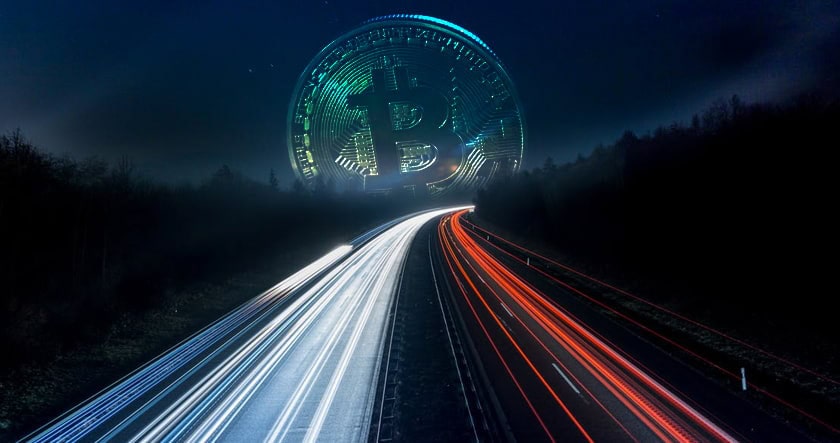 A large digital Bitcoin coin appears in the sky above a highway at night, with car lights creating long streaks along the road.