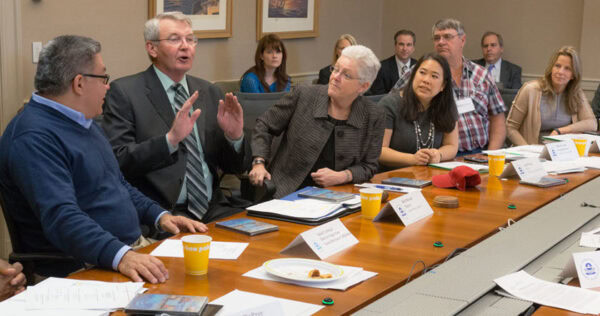 Several people sit around a conference table engaged in discussion, with papers, name tags, and coffee cups visible on the table.