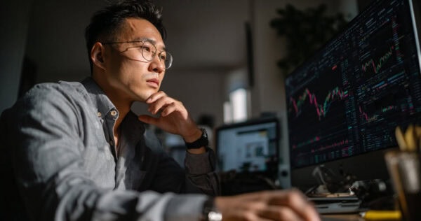 A man wearing glasses sits at a desk, looking at multiple computer monitors displaying financial charts and data.