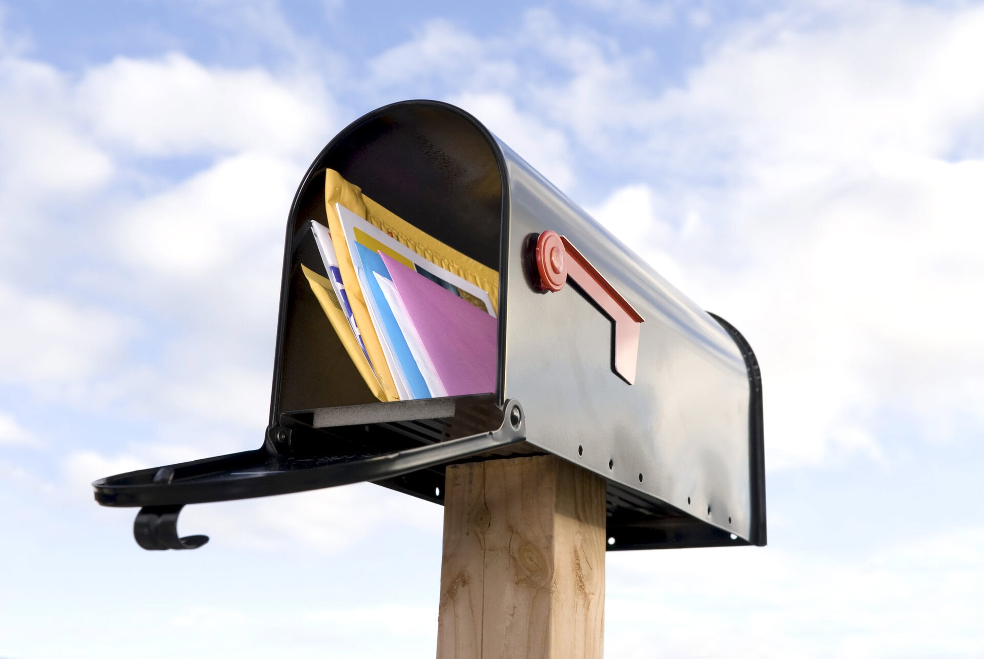 A black mailbox with its door open, mounted on a wooden post, is filled with colorful envelopes and packages. The background shows a blue sky with scattered white clouds.