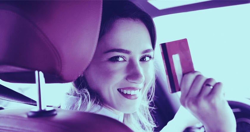 A woman sitting in a car holds up a credit card and smiles at the camera.