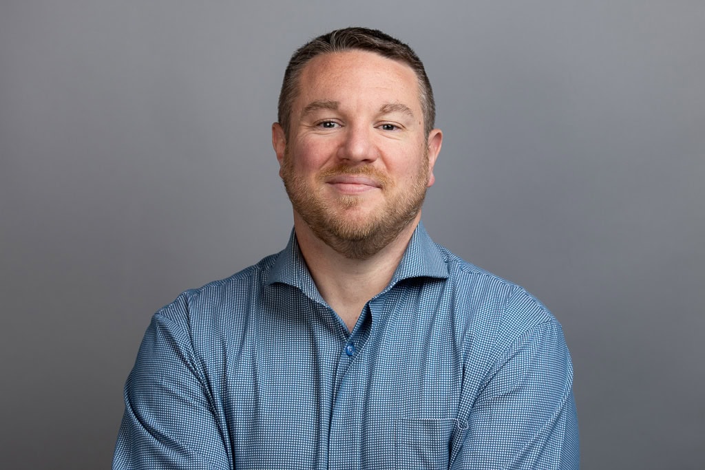 A man with short hair and a trimmed beard wearing a blue checkered shirt stands against a plain gray background, looking at the camera with a slight smile and arms crossed.